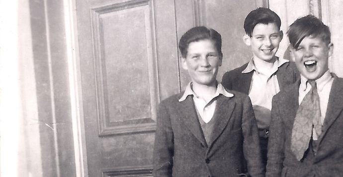 Three boys in suits stand smiling in front of a wooden door, with one boy laughing energetically.