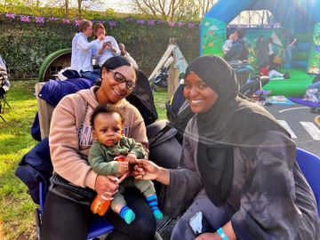 A woman holds a baby on her lap while another woman smiles beside them at an outdoor event with a bouncy castle in the background.
