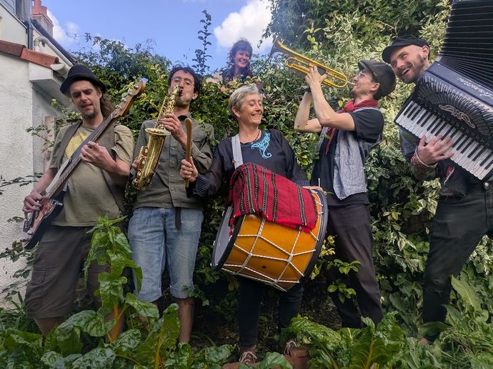 A group of musicians performing outdoors with guitar, saxophone, trumpet, drum, and accordion, surrounded by lush greenery and a clear sky.