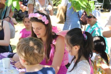 A group of children gathers around a person dressed in a pink costume with flower accessories at an outdoor activity table.