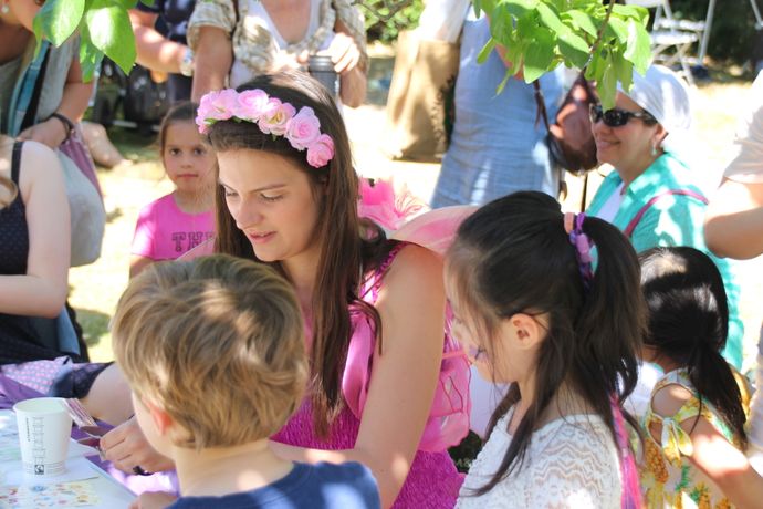 A group of children gathers around a person dressed in a pink costume with flower accessories at an outdoor activity table.