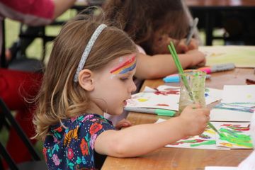 A child sits at a table using a paintbrush to create a colourful artwork among scattered art supplies.