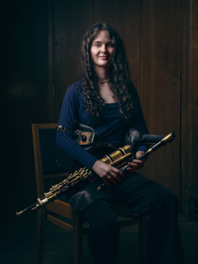 Young woman with long curly hair sits on a chair, holding a set of uilleann pipes, wearing a dark blue top and black pants, in a dimly lit room.
