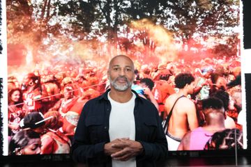 A person stands in front of a vibrant photo of a lively festival crowd covered in red powder, surrounded by trees.
