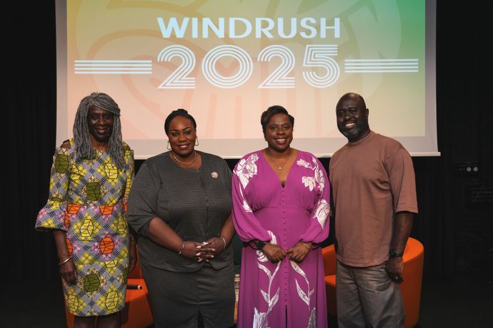 Four people stand smiling in front of a "Windrush 2025" sign. They are dressed in colorful and formal attire.