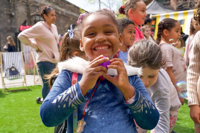 A child standing on grass holds a small object while surrounded by other children at an outdoor event with bright decorations.