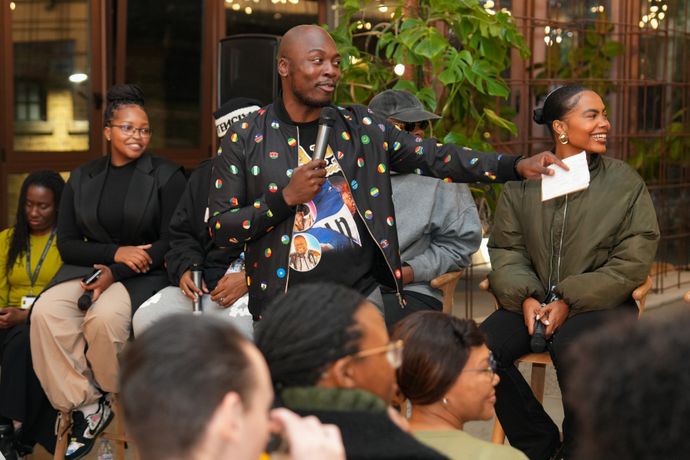 A man speaks into a microphone, gesturing towards a smiling woman. Several people are seated around them in a lively indoor setting.