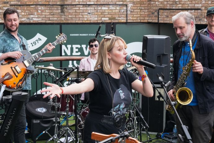 A band of individuals playing instruments with the lead singer wearing an Amy Winehouse top