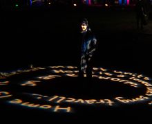 Person standing on a dark path illuminated by circular projected text, wearing a coat and scarf, with blurred lights in the background.