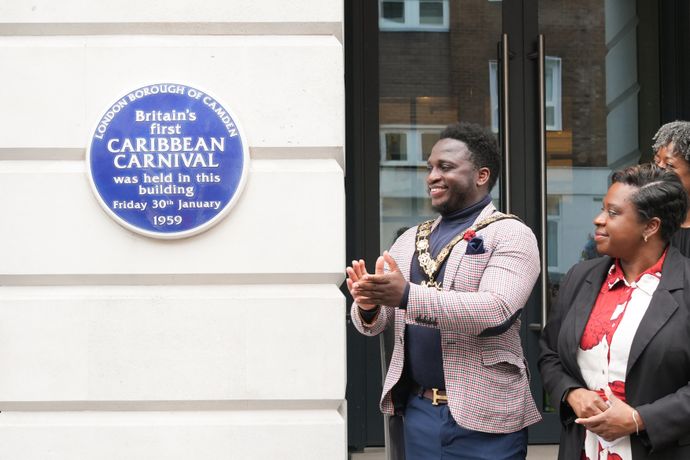 People standing near a blue plaque commemorating Britain's first Caribbean Carnival in Camden, dated January 30, 1959.