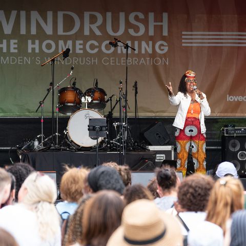 Performer on stage at Windrush Homecoming event, addressing a diverse audience. Drums and microphones are visible in the background.
