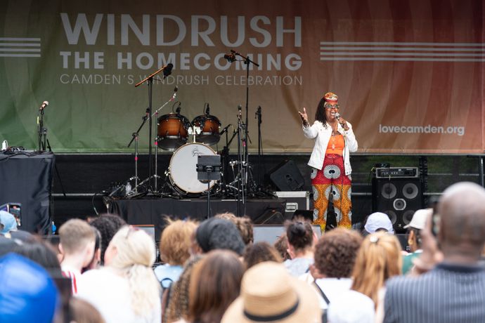 Performer on stage at Windrush Homecoming event, addressing a diverse audience. Drums and microphones are visible in the background.