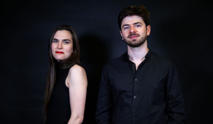 Two people standing against a dark background, both wearing black clothing.