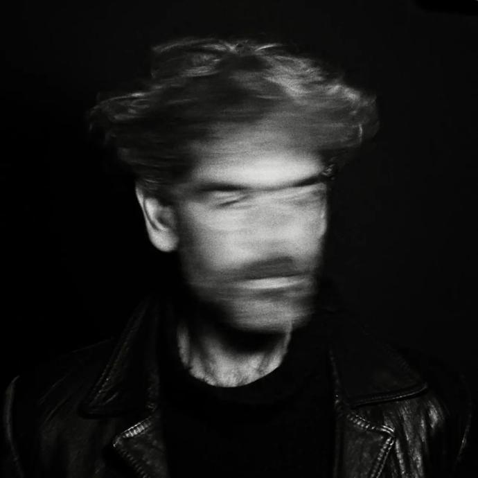 A black and white image of a man with blurred face motion, wearing a leather jacket, set against a dark background.