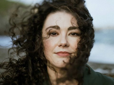 A woman with curly hair stands outdoors near a beach, with soft waves and greenery in the background. Her hair is blowing gently in the wind.