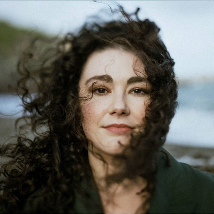 A woman with curly hair stands outdoors near a beach, with soft waves and greenery in the background. Her hair is blowing gently in the wind.