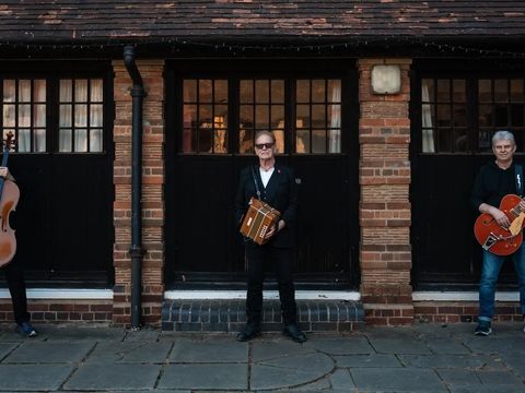 Three musicians stand in front of an old brick building, holding a cello, accordion, and electric guitar.