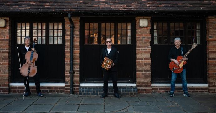 Three musicians stand in front of an old brick building, holding a cello, accordion, and electric guitar.