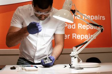 A technician wearing gloves and goggles repairs a smartphone on a workbench under a desk lamp, with repair tools scattered around.