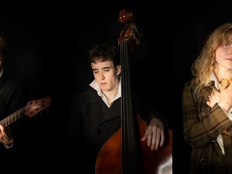 Three people in dark jackets stand against a black background; one plays an electric guitar, one plays a double bass, and one holds a closed book to their chest.