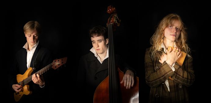 Three people in dark jackets stand against a black background; one plays an electric guitar, one plays a double bass, and one holds a closed book to their chest.