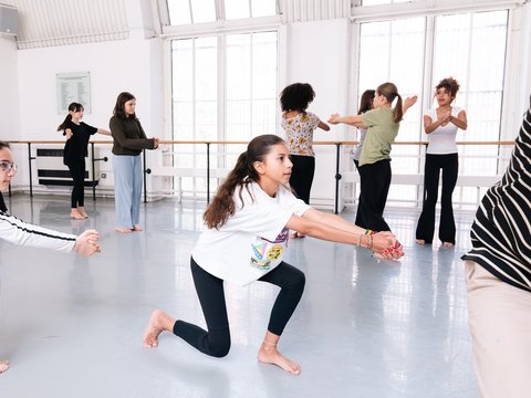 A diverse group of people practice dance movements in a bright studio, with one girl in the foreground stretching her arms forward.