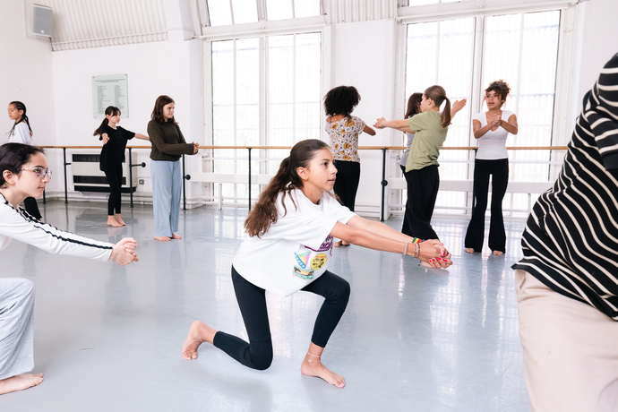 A diverse group of people practice dance movements in a bright studio, with one girl in the foreground stretching her arms forward.