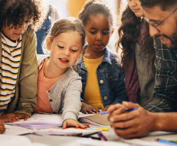four children crowd around a table with paper and pens, one reaches across the paper as an adult leans over the table watching and talking