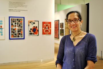 Smiling woman in a blue shirt stands in a gallery with colorful abstract artwork on the wall behind her.