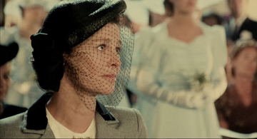Woman in a gray suit and hat with a veil looks to the side, standing in a crowd. A person in white attire is blurred in the background.