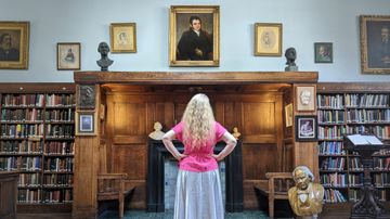 A person stands in a wood‑panelled library, facing a wall of portraits and bookshelves with hands on hips.