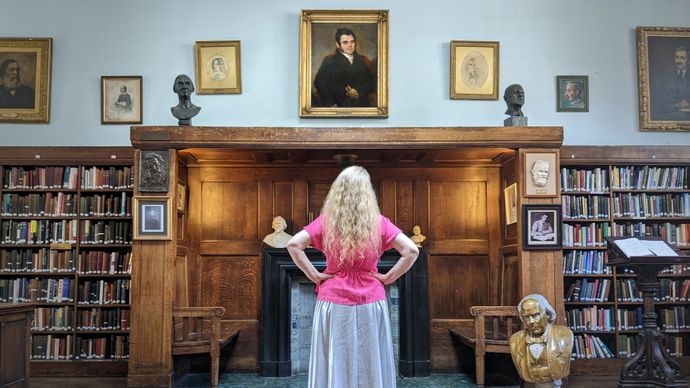 A person stands in a wood‑panelled library, facing a wall of portraits and bookshelves with hands on hips.