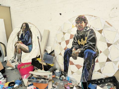 Two mosaic artworks on circular panels depict people sitting, surrounded by art materials and crates in a workshop setting.