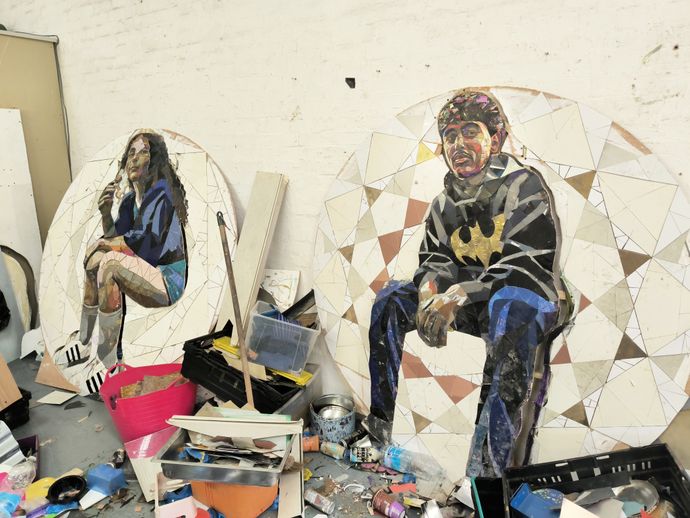Two mosaic artworks on circular panels depict people sitting, surrounded by art materials and crates in a workshop setting.