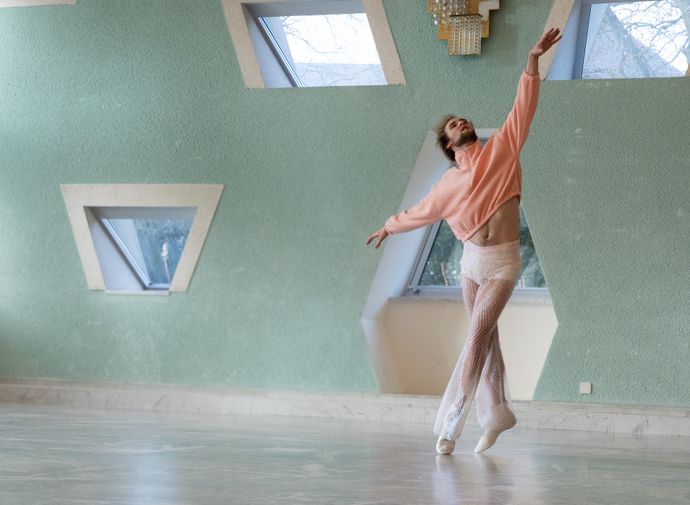 A dancer in a pink sweater and sheer pants strikes an elegant pose in a studio with mint green walls and triangular windows.