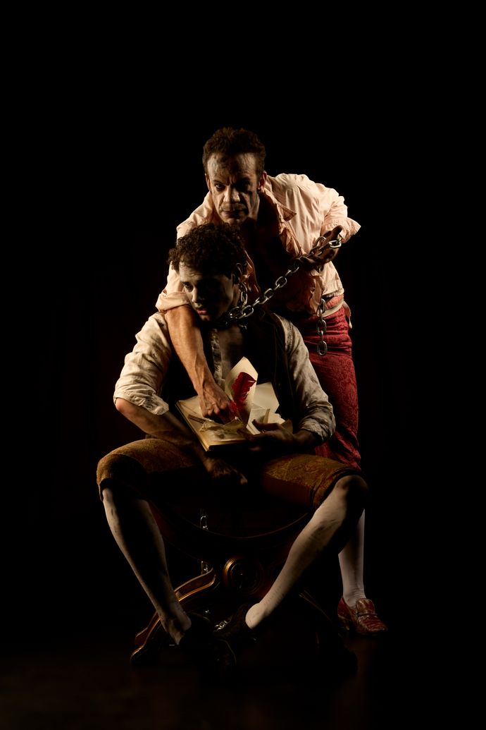 Two men on stage, one seated holding a book, the other standing with chains, in dramatic lighting and theatrical setting.