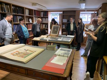 A group of people in a library examines artwork and documents displayed on tables, surrounded by bookshelves.