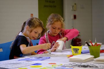 Two young girls are focused on arts and crafts, working intently at a table filled with paint and paper, both wearing aprons.