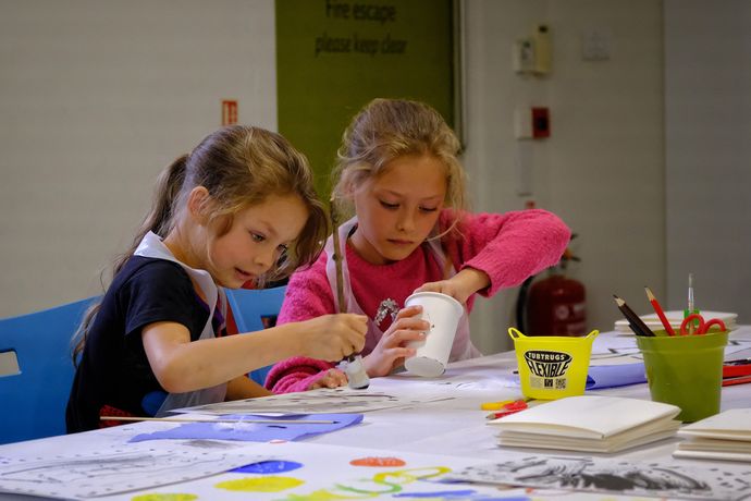 Two young girls are focused on arts and crafts, working intently at a table filled with paint and paper, both wearing aprons.