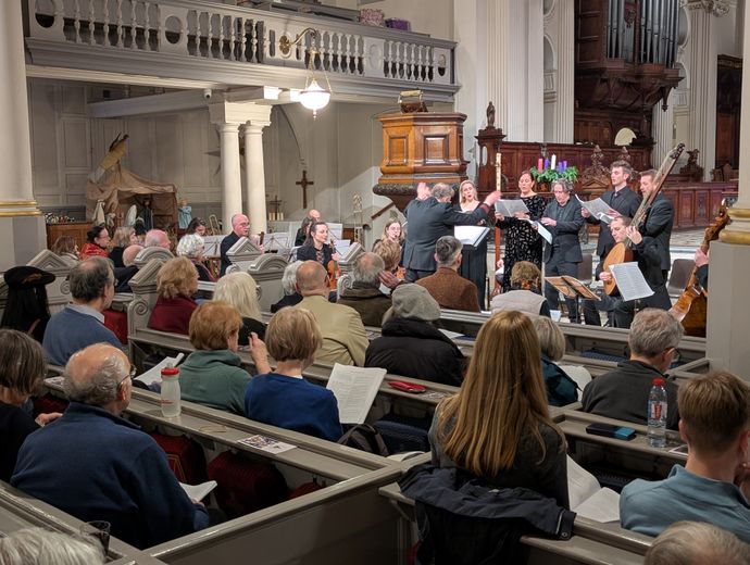 Indoor concert in a historic church with musicians performing at the front, holding sheet music and playing instruments, while an audience sits in pews following along with programs. Visible features include wooden pulpit, organ pipes, and a nativity scene in the background.