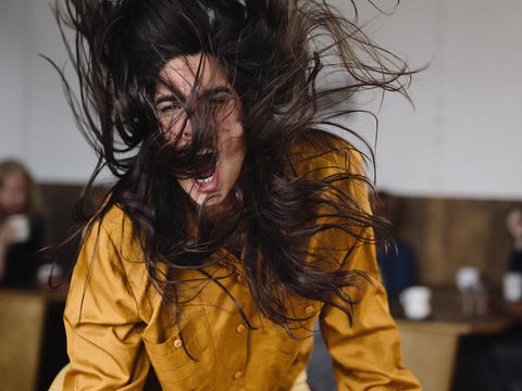 Woman with long dark brown hair slams fist with force wearing yellow mustard shirt, hair is in wild movement with the force of movement