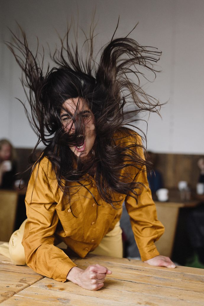 Woman with long dark brown hair slams fist with force wearing yellow mustard shirt, hair is in wild movement with the force of movement
