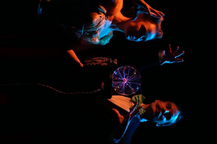 A person in formal attire and another in a costume pose dramatically in dim lighting around a glowing plasma ball, creating a theatrical and mysterious scene.