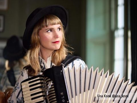 Person wearing a dark hat plays accordion indoors, beside window, with blurred reflection behind.