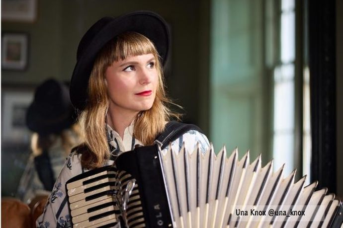 Person wearing a dark hat plays accordion indoors, beside window, with blurred reflection behind.