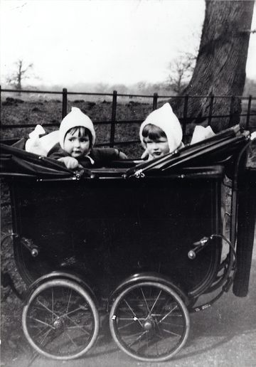 Two young children wearing white bonnets sit in a vintage double pram outdoors, with a tree and fence in the background.
