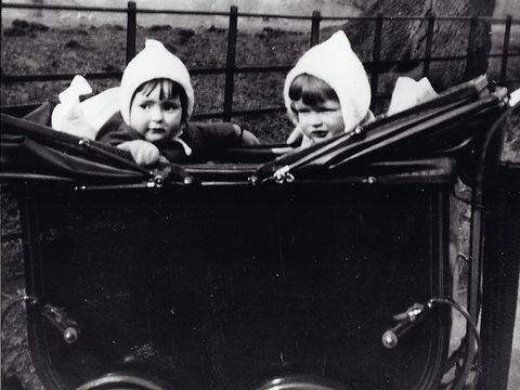 Two young children wearing white bonnets sit in a vintage double pram outdoors, with a tree and fence in the background.