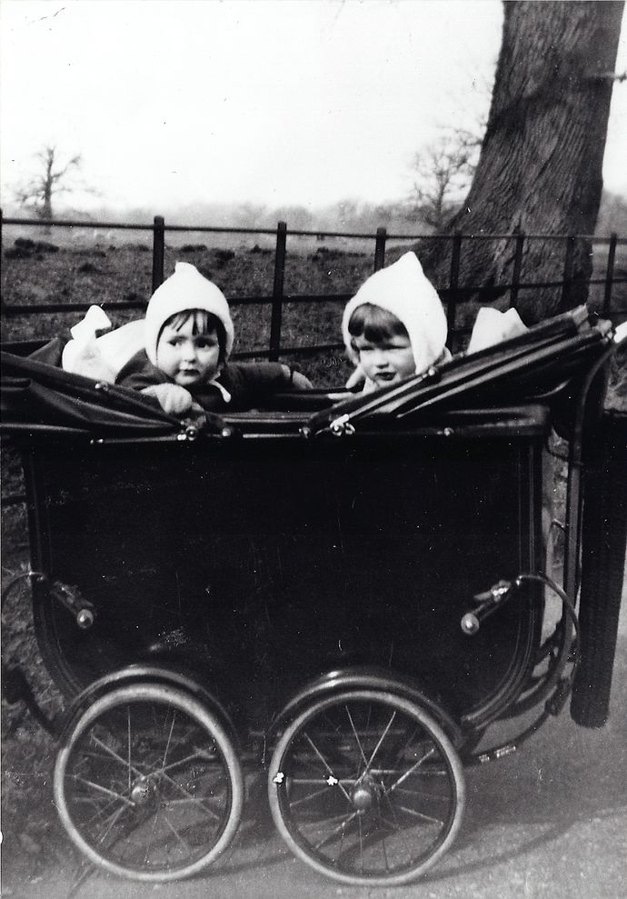 Two young children wearing white bonnets sit in a vintage double pram outdoors, with a tree and fence in the background.