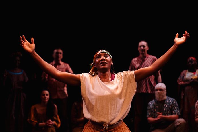 A person on stage extends their arms joyfully, with several people in the background, under warm spotlighting.