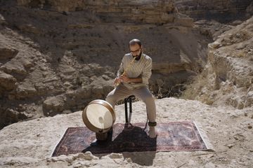 A person seated on a stool outdoors in a rocky canyon, playing a wooden wind instrument. A large frame drum rests on a patterned rug beneath them. The scene is sunlit, with rugged cliffs in the background.Provide your feedback on BizChat